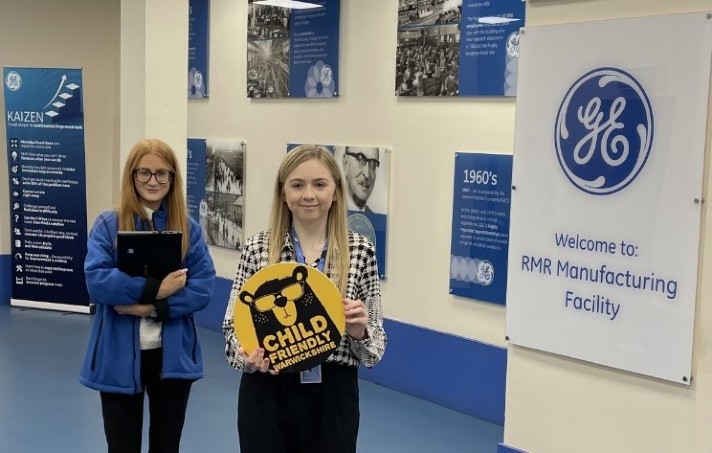 Two young girls stood holing the Child Friendly Warwickshire logo at the General Electric facility.